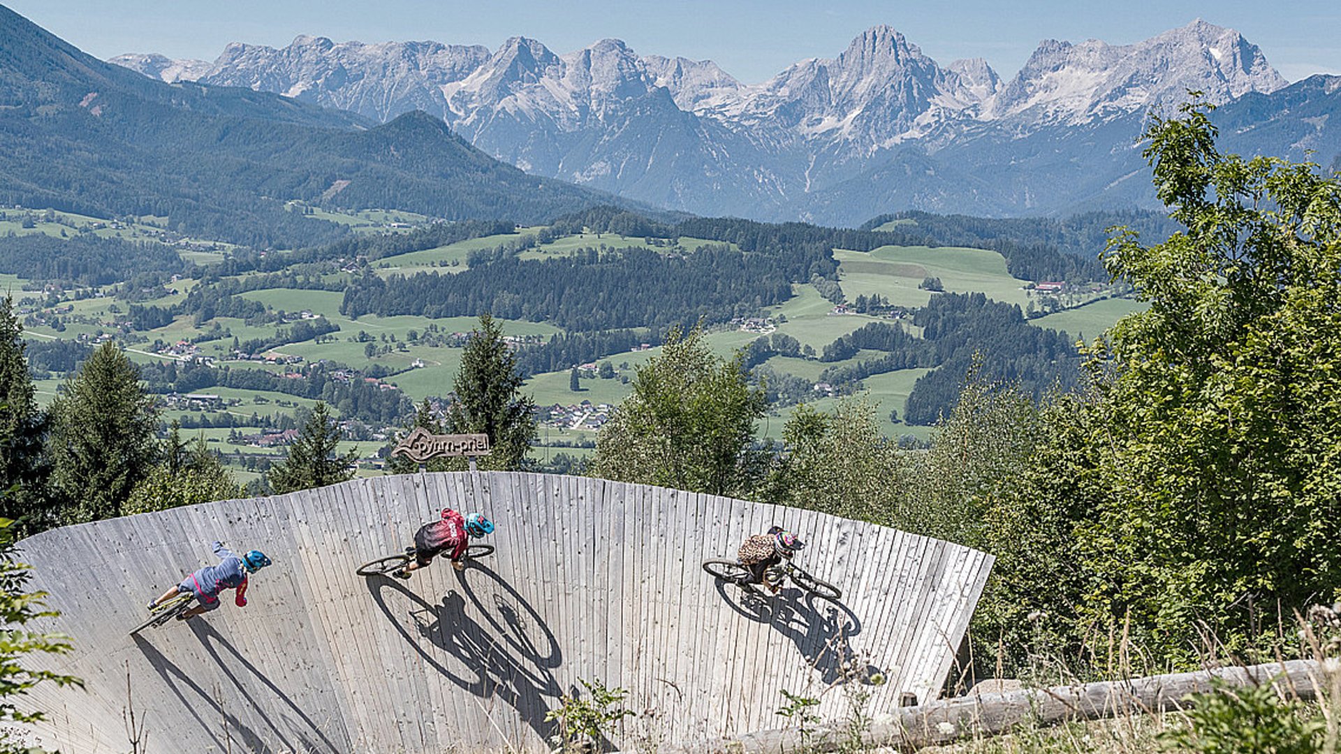 Foto Oberösterreich Tourismus GmbH/Daniel Hinterramskolger: Drei Biker in der Steilkurve des Bikeparks Wurbauerkogel in Windischgarsten, im Hintergrund das Tote Gebirge. Drei Biker in der Steilkurve des Bikeparks Wurbauerkogel in Windischgarsten, im Hintergrund das Tote Gebirge.